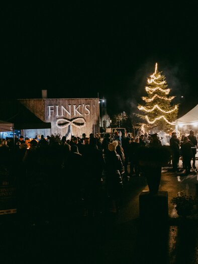 Eine festliche Veranstaltung bei Nacht mit vielen Menschen. Ein geschmückter Weihnachtsbaum leuchtet vor dem Gebäude von FINK'S. | © Haberl Fink