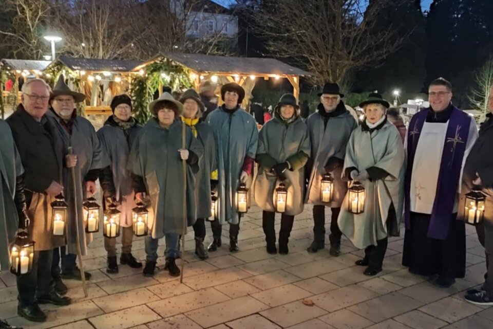 Eine Gruppe von Menschen in grauen Umhängen steht draußen bei Nacht. Sie halten Laternen und lächeln, während im Hintergrund festliche Lichter zu sehen sind. | © Gemeinde Bad Gleichenberg