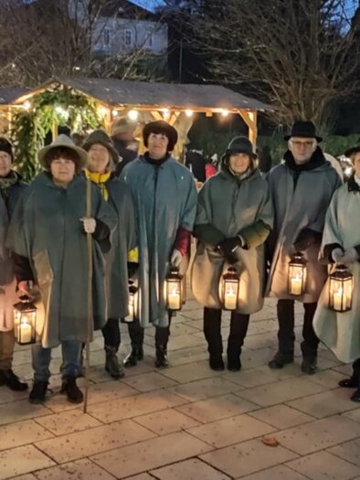Eine Gruppe von Menschen in grauen Umhängen steht draußen bei Nacht. Sie halten Laternen und lächeln, während im Hintergrund festliche Lichter zu sehen sind. | © Gemeinde Bad Gleichenberg