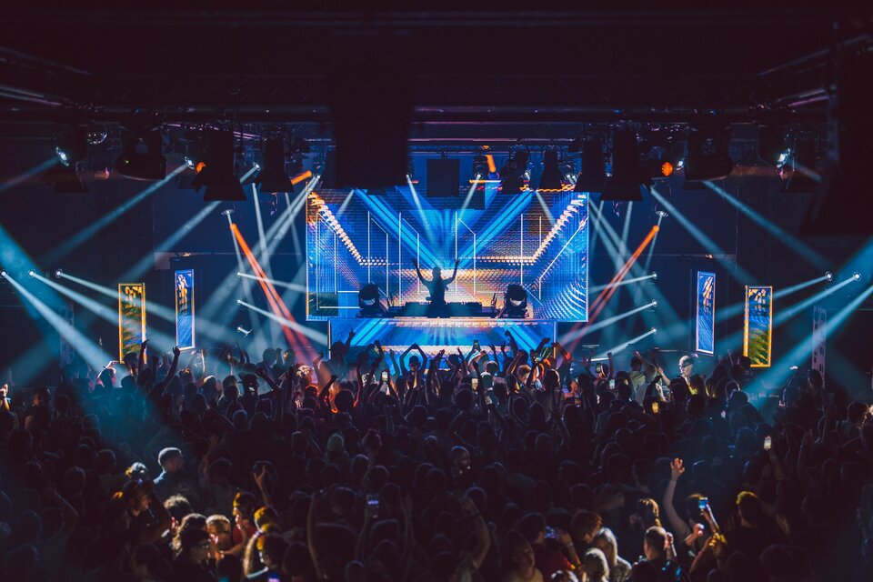 Eine energiegeladene Live-Performance auf einer Bühne mit bunten Lichtern. Das Publikum ist begeistert und tanzt zur Musik. | © Luke Goodlife