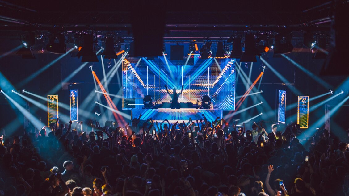 Eine energiegeladene Live-Performance auf einer Bühne mit bunten Lichtern. Das Publikum ist begeistert und tanzt zur Musik. | © Luke Goodlife