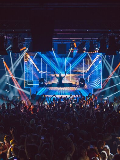 Eine energiegeladene Live-Performance auf einer Bühne mit bunten Lichtern. Das Publikum ist begeistert und tanzt zur Musik. | © Luke Goodlife
