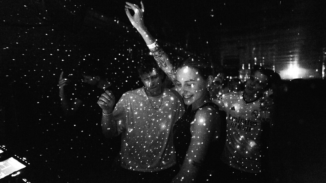 Eine Gruppe von Menschen tanzt in einem dunklen Raum, der von funkelnden Lichtern erleuchtet wird. Die Stimmung ist lebhaft und fröhlich. | © Stefan Leitner