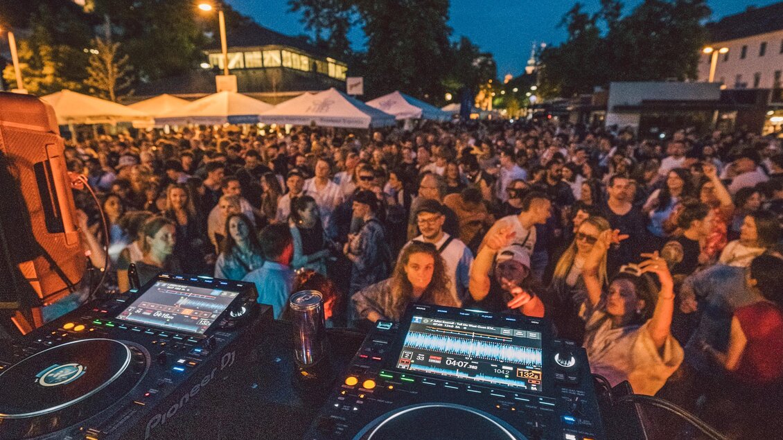 Eine lebhafte Konzertatmosphäre mit einer großen Menschenmenge unter einem klaren Abendhimmel. Im Vordergrund sind DJ-Pulte sichtbar, die zum Feiern einladen. | © Luke Goodlife