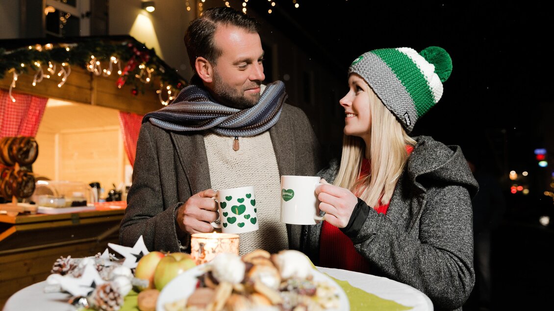 Ein Paar steht an einem Tisch und hält Tassen in der Hand. Im Hintergrund sind festliche Lichter und ein Weihnachtsstand zu sehen. | ©  Schilcherland Steiermark - Lupi Spum