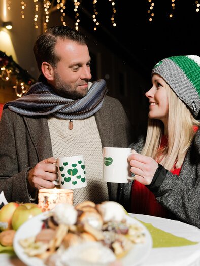 A couple is standing at a table holding cups in their hands. In the background, festive lights and a Christmas stall can be seen. | ©  Schilcherland Steiermark - Lupi Spum