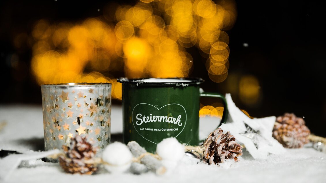 Ein grüner Becher steht neben einem glitzernden Teelicht auf einer schneebedeckten Oberfläche. Im Hintergrund sind weiche Lichter zu sehen, die eine festliche Stimmung erzeugen. | ©  Lupi Spuma