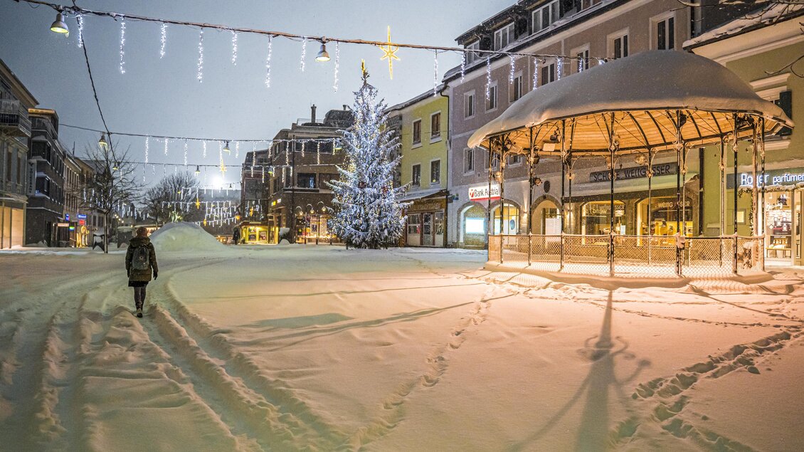 Eine verschneite Stadtmitte in der Nacht mit einem festlich beleuchteten Baum. Eine Person geht durch den dicken Schnee, während die Gebäude im Hintergrund sichtbar sind. | © Gerhard Pilz
