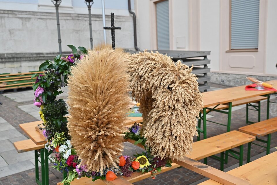 Ein dekorativer Wreath aus trockenem Gras und bunten Blumen steht auf einem Tisch. Im Hintergrund sind Bänke und eine Wand zu sehen. | © Norbert Ortner