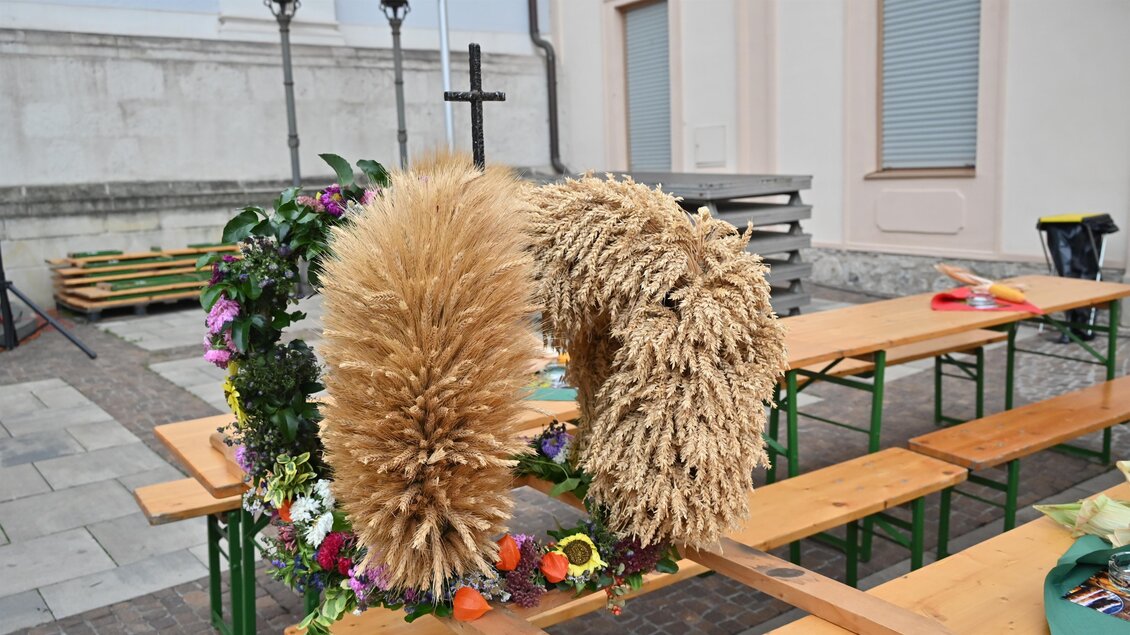Ein dekorativer Wreath aus trockenem Gras und bunten Blumen steht auf einem Tisch. Im Hintergrund sind Bänke und eine Wand zu sehen. | © Norbert Ortner
