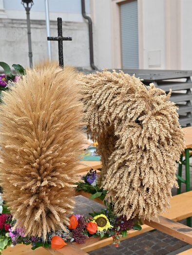 A decorative wreath made of dried grass and colorful flowers is on a table. In the background, benches and a wall can be seen. | © Norbert Ortner