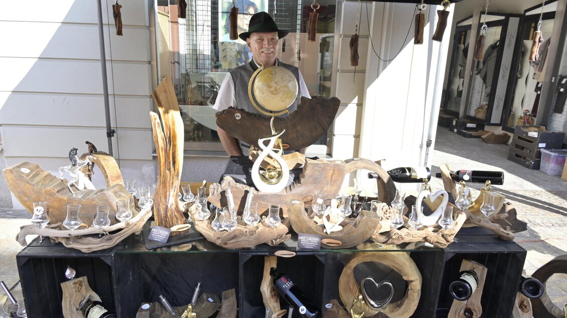 Ein Marktstand mit handgefertigten Holzgegenständen und Schmuck. Im Hintergrund steht ein Verkäufer, der die Waren präsentiert. | © Norbert Ortner