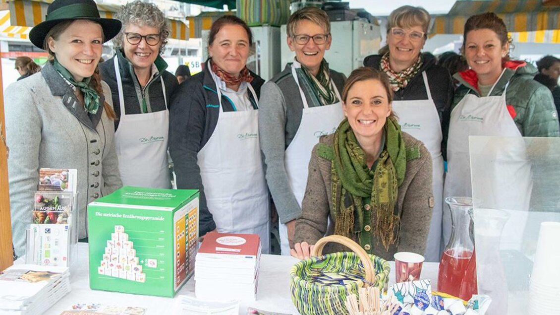Eine Gruppe von Frauen steht lächelnd an einem Marktstand. Sie tragen Schürzen und präsentieren lokale Produkte. | © Pressberger