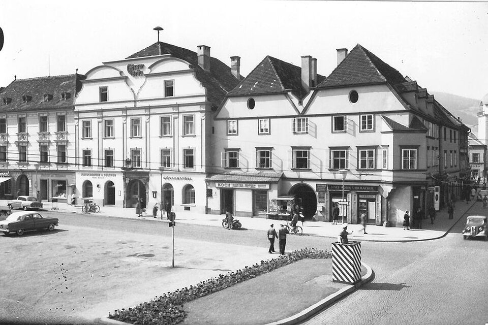 Ein historischer Stadtplatz mit alten Gebäuden und einem kleinen Platz. Fahrzeuge und Passanten sind sichtbar, was das lebendige Flair der Szene unterstreicht. | ©  MuseumsCenter Leoben