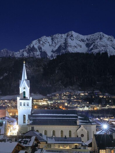 A picturesque town at night, surrounded by snow-covered mountains. The churches with their characteristic towers shine in the light of the city. | © Martin Huber