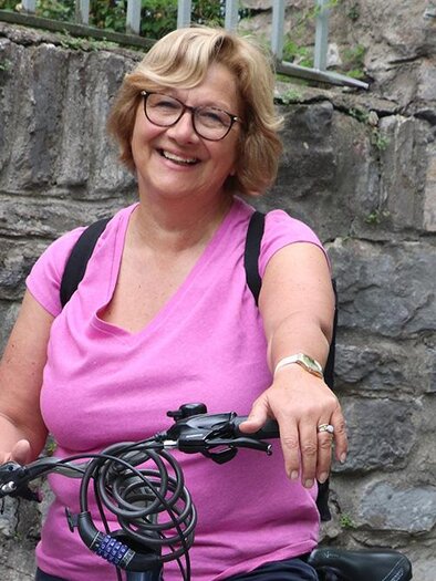 A smiling woman with glasses is sitting on a bicycle. She is wearing a pink T-shirt and has a backpack-like item with her. | ©  leopress