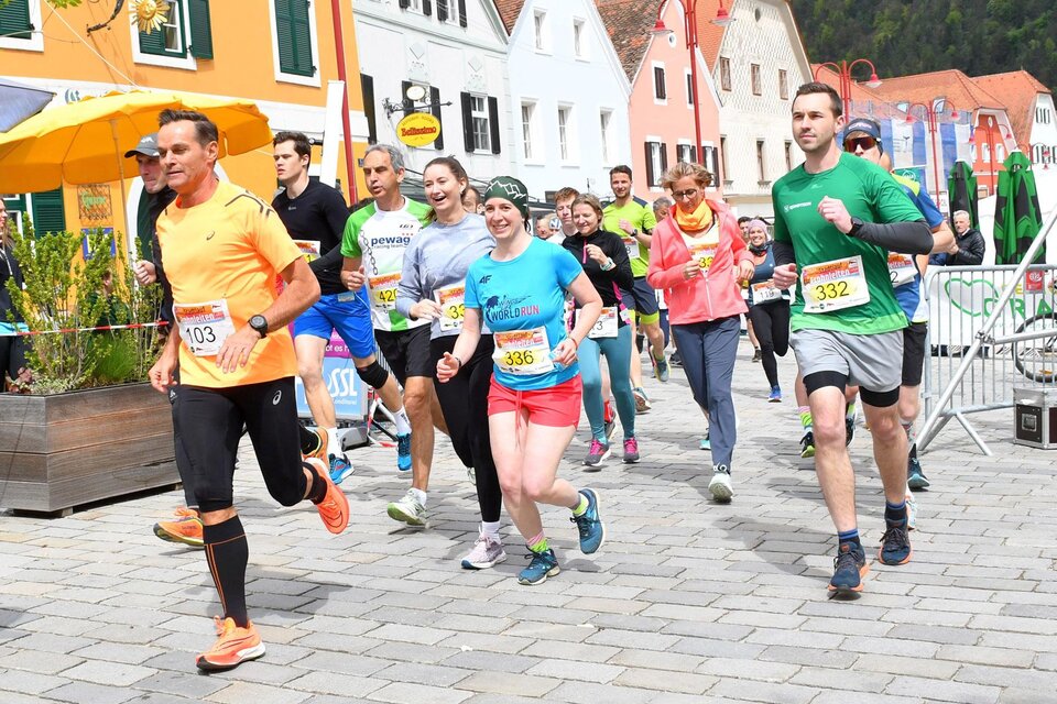 Eine Gruppe von Läufern nimmt an einem Wettkampf in einer malerischen Stadt teil. Sie tragen bunte Sportkleidung und lächeln, während sie die Straße entlanglaufen. | © WOKA