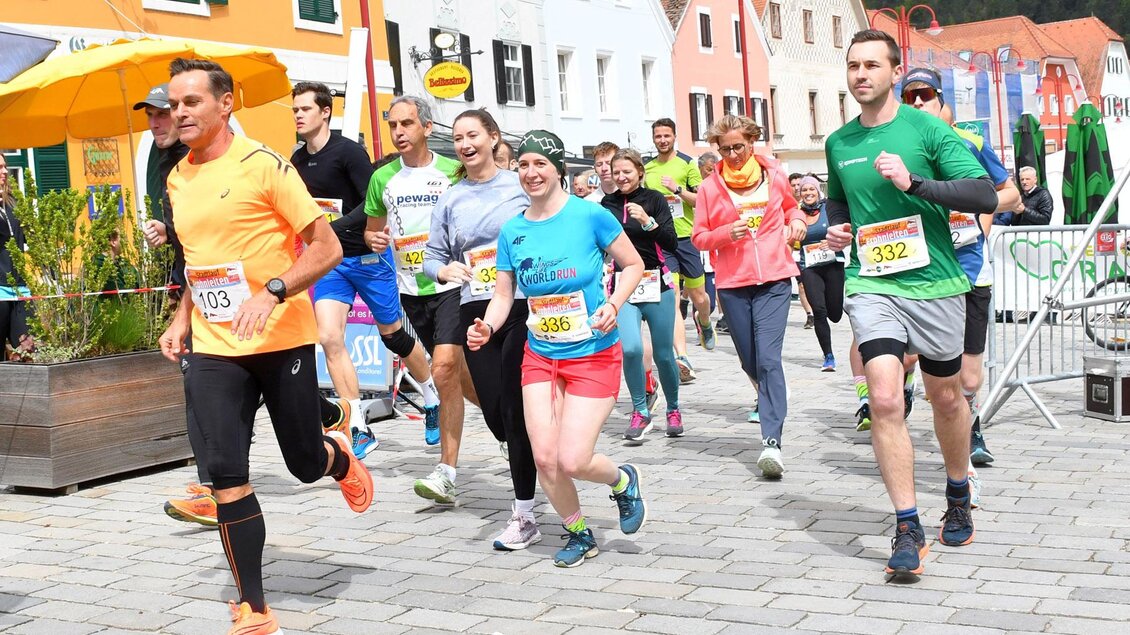 Eine Gruppe von Läufern nimmt an einem Wettkampf in einer malerischen Stadt teil. Sie tragen bunte Sportkleidung und lächeln, während sie die Straße entlanglaufen. | © WOKA
