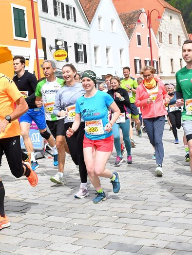 A group of runners is participating in a competition in a picturesque town. They are wearing colorful sportswear and smiling as they run along the street. | © WOKA