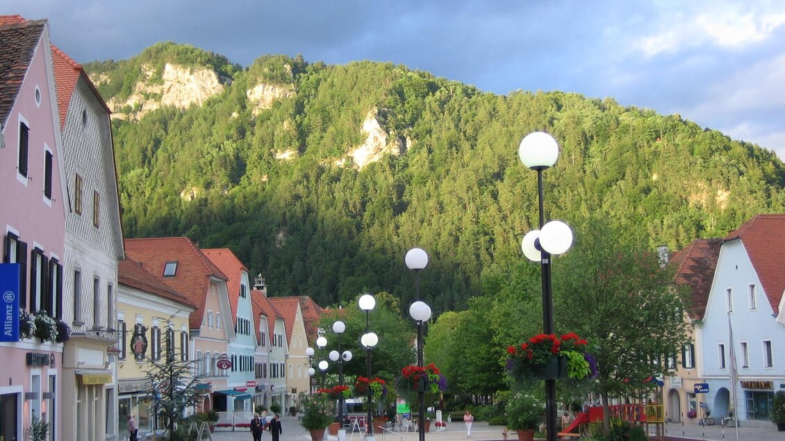 Eine malerische Straße mit bunten Häusern und Laternen. Im Hintergrund erhebt sich ein bewaldeter Berg. | © TV Frohnleiten-Michael Dittrich
