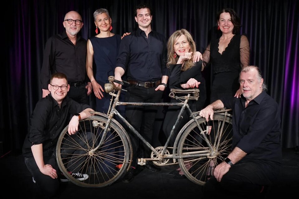 Eine Gruppe von sechs Personen posiert um ein altes Fahrrad. Sie tragen elegante schwarze Kleidung und lächeln in die Kamera. | © StainZeit - Theatertruppe Johann Fink