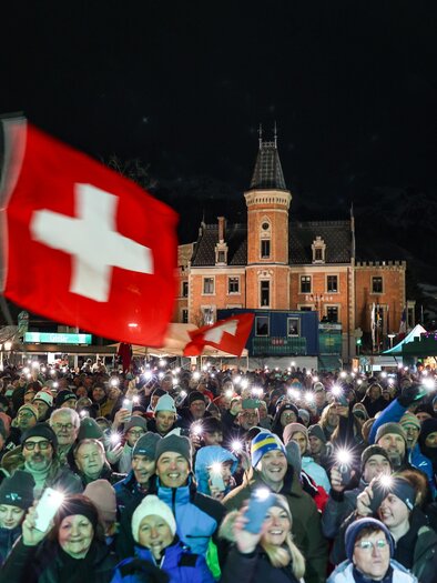 A large crowd is holding mobile lights in the air and waving flags. In the background, a historic building can be seen at night. | © Martin Huber
