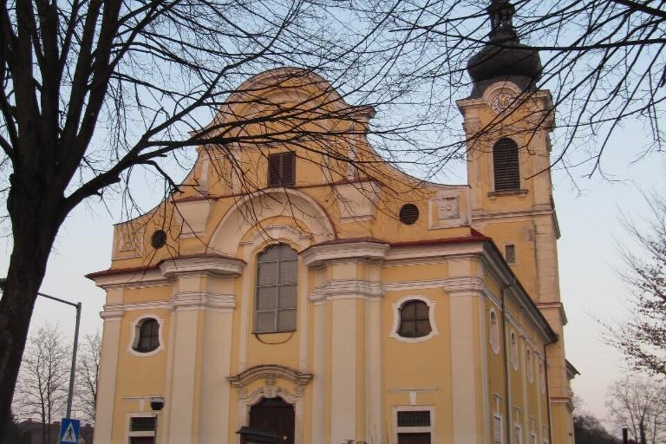 Eine gelbe Kirche mit einem hohen Turm und schönen architektonischen Details. Im Hintergrund sind kahler Baum und der Abendhimmel zu sehen.