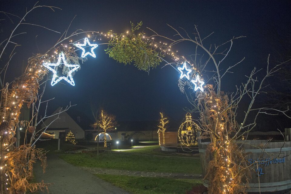 Ein festlicher Lichtbogen mit leuchtenden Sternen und grünen Ästen erhellt die Nacht. Im Hintergrund sind weitere dekorative Lichter und eine ruhige Landschaft zu sehen. | © Gemeinde Bad Loipersdorf