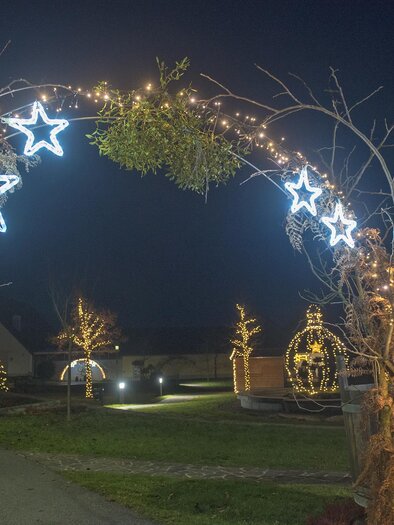 A festive light arch with shining stars and green branches illuminates the night. In the background, more decorative lights and a tranquil landscape can be seen. | © Gemeinde Bad Loipersdorf