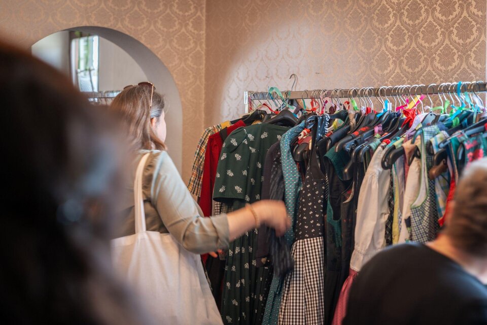 Eine Frau schaut sich Kleidungsstücke an einem Kleiderständer an. Der Hintergrund zeigt eine stilvolle Wandgestaltung. | © Haus der Frauen