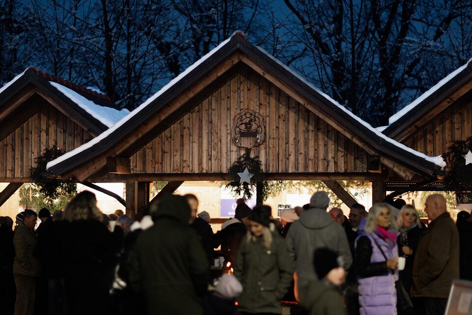 Ein belebter Weihnachtsmarkt mit festlich dekorierten Holzhütten. Menschen genießen die Atmosphäre in der Abenddämmerung. | ©  Achromatic Photography