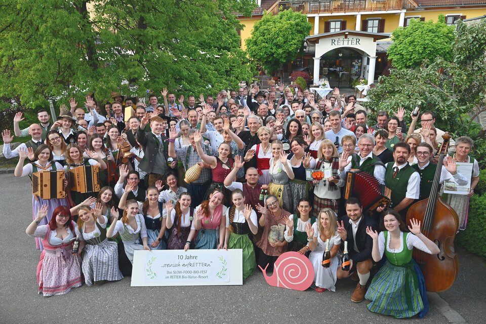 Eine große Gruppe von Menschen in traditioneller Kleidung posiert fröhlich im Freien. Im Hintergrund sind Bäume und ein Gebäude sichtbar. | © RETTER- BIO- Natur-Resort