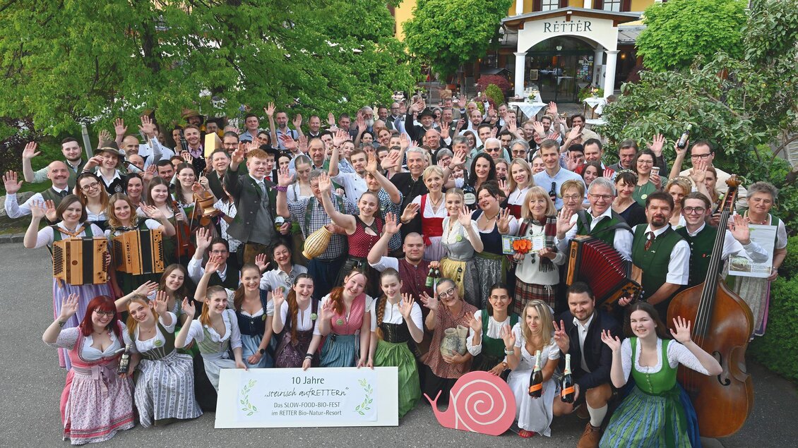 Eine große Gruppe von Menschen in traditioneller Kleidung posiert fröhlich im Freien. Im Hintergrund sind Bäume und ein Gebäude sichtbar. | © RETTER- BIO- Natur-Resort