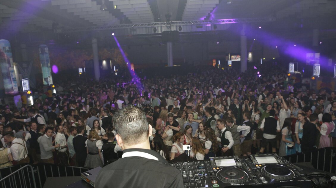 Eine große Menge von Menschen tanzt und feiert in einem Veranstaltungsraum. Im Vordergrund steht ein DJ, der Musik auflegt. | © Arthur
