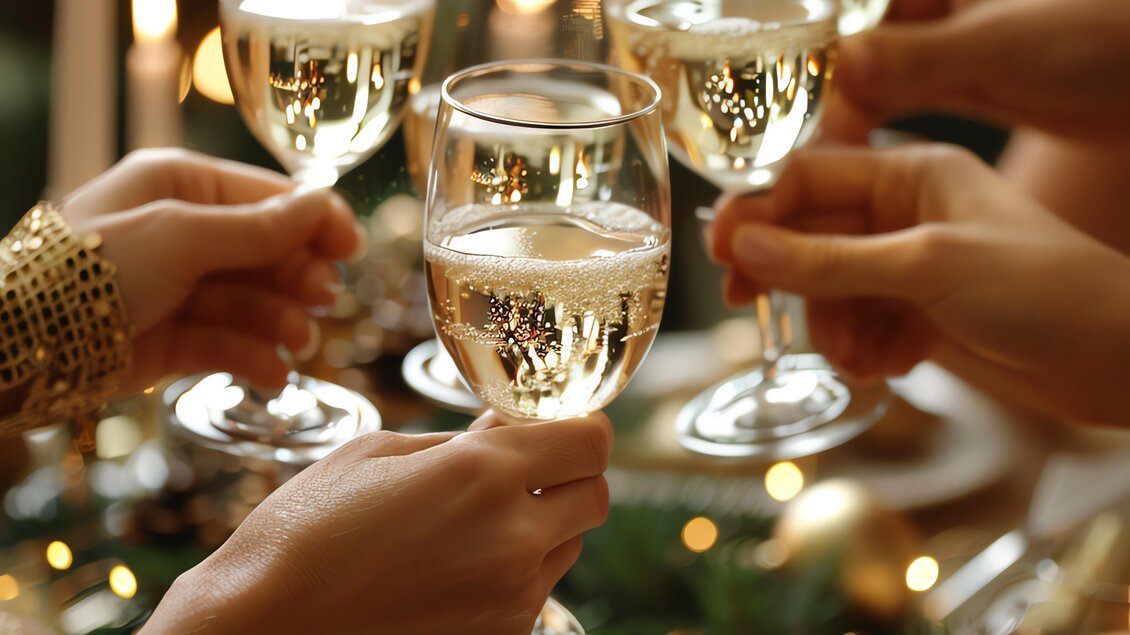 Ein festlicher Anstoß mit Sektgläsern in einer gemütlichen Atmosphäre. Im Hintergrund leuchten Kerzen und Weihnachtsdekoration. | © MALU