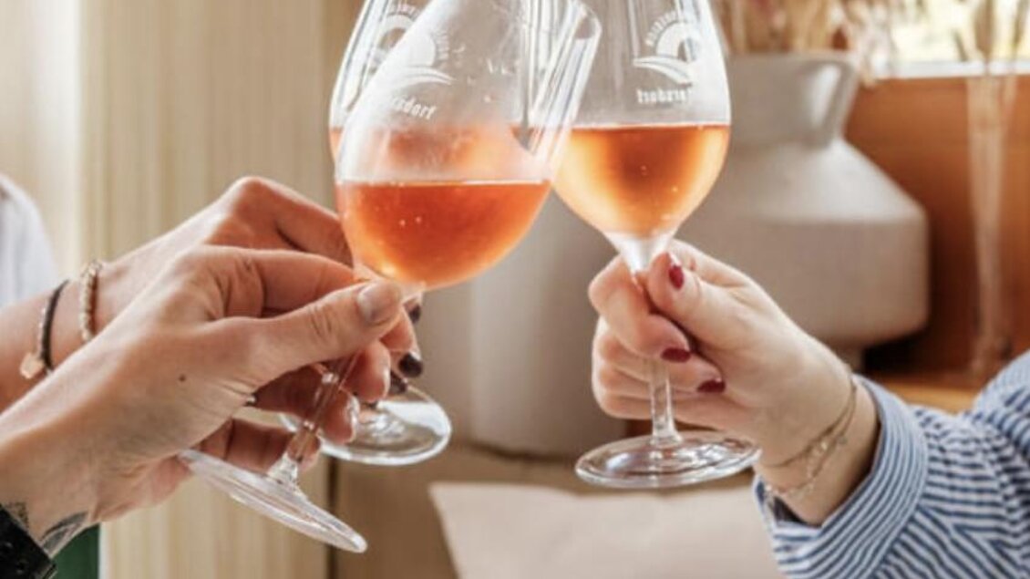 Ein fröhliches Anstoßen mit Gläsern voller Roséwein. Im Hintergrund sind festliche Dekorationen und ein gemütliches Ambiente zu sehen. | © Malu