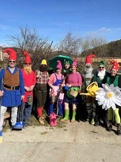 A group of people in colorful garden gnome costumes is happily standing together. In the background, there are trees and a blue sky visible. | © Stern-Thaler Laientheater