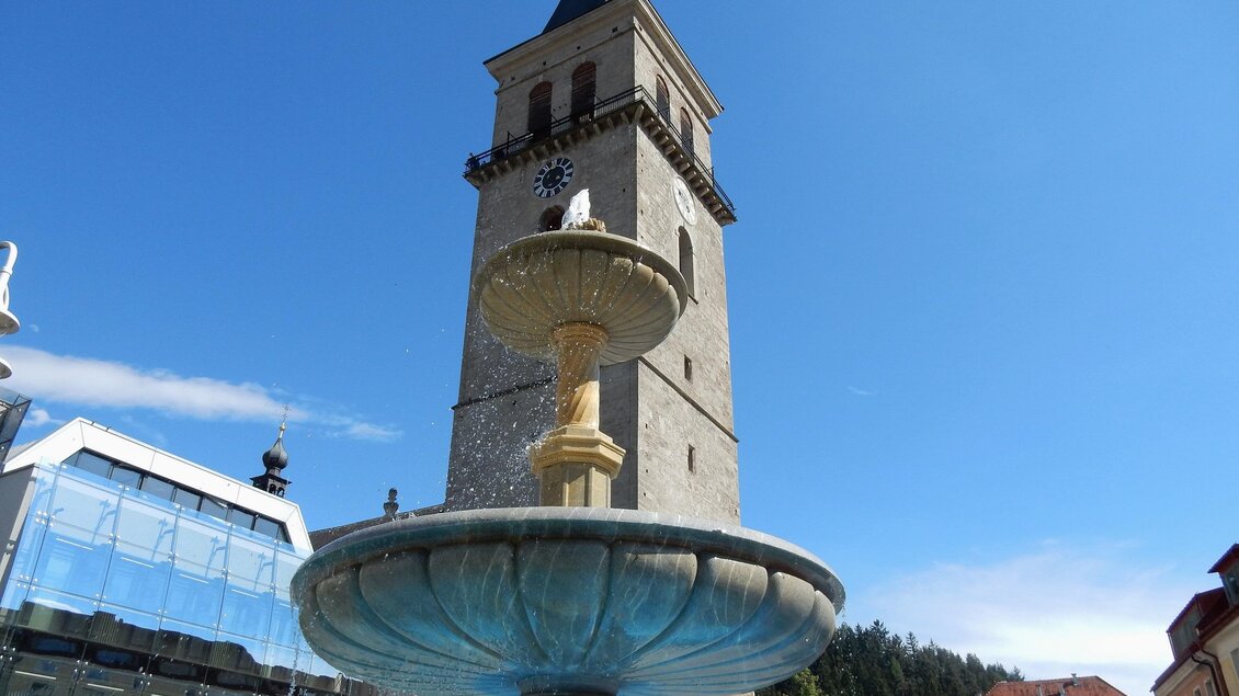 Ein hübscher Brunnen mit einer Wasserfontäne im Vordergrund. Im Hintergrund steht ein alter Uhrturm unter klarem, blauem Himmel. | © Sternenturm Judenburg