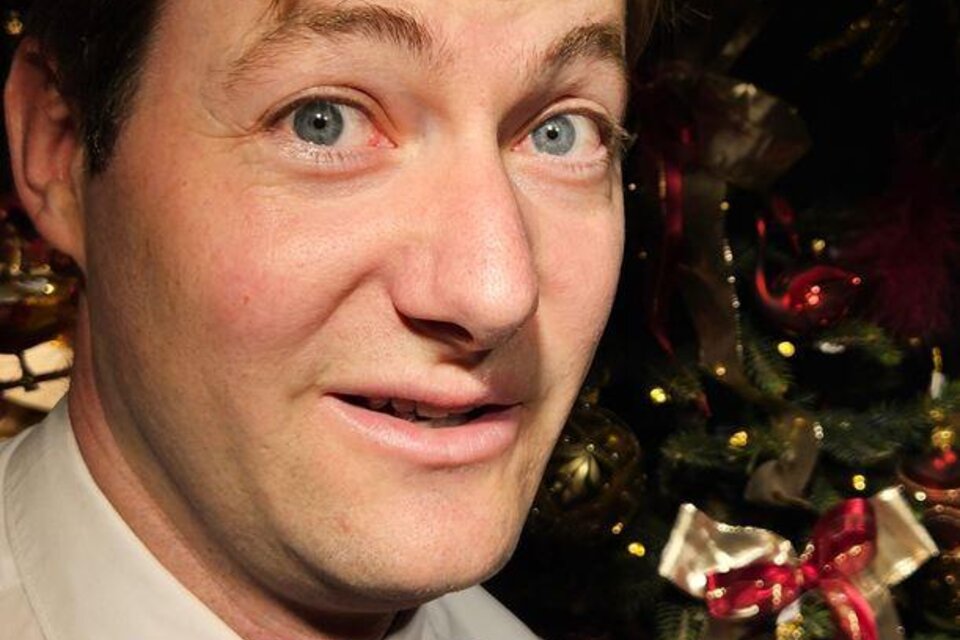 Porträt eines Mannes mit weißem Hemd und roter Fliege, der leicht lächelnd in die Kamera blickt; im Hintergrund weihnachtlich geschmückter Baum mit roten und goldenen Ornamenten. | © Großschädl