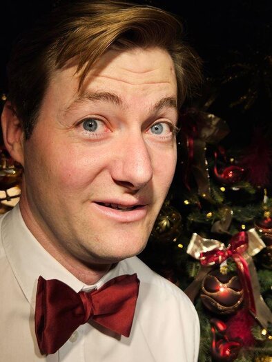 Portrait of a man in a white shirt and red bow tie, slightly smiling at the camera; in the background, a Christmas tree decorated with red and gold ornaments. | © Großschädl