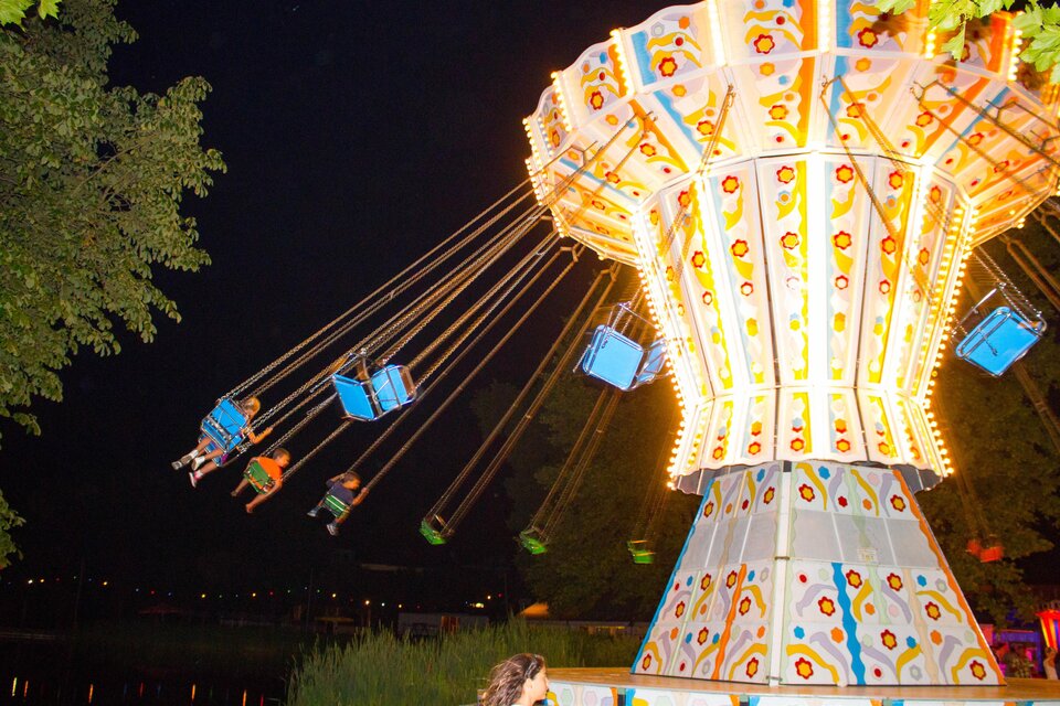 Eine bunte Karussellfahrt mit leuchtenden Lichtern, die Kinder hoch in die Luft schwingt. Der Hintergrund ist abends mit einem ruhigen See zu sehen. | © Sandra Brünner