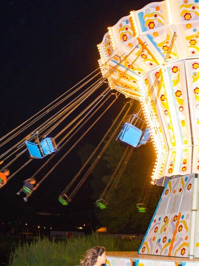A colorful carousel ride with bright lights that swings children high into the air. The background features a calm lake in the evening. | © Sandra Brünner