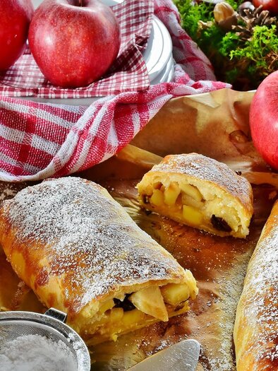In the picture lies a freshly baked, golden-brown apple strudel, dusted with powdered sugar. All around are red apples and a red-and-white checkered cloth. | © Pixabay