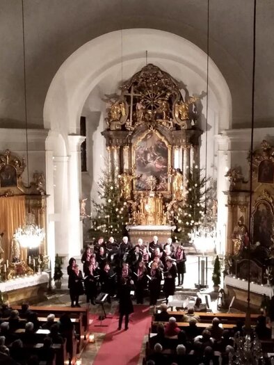 Ein festlicher Chor tritt in einer schönen Kirche auf. Die dekorierte Altaranlage und die mit Lichtern geschmückten Bäume sorgen für eine warme Atmosphäre. | © Singkreis Stubenberg