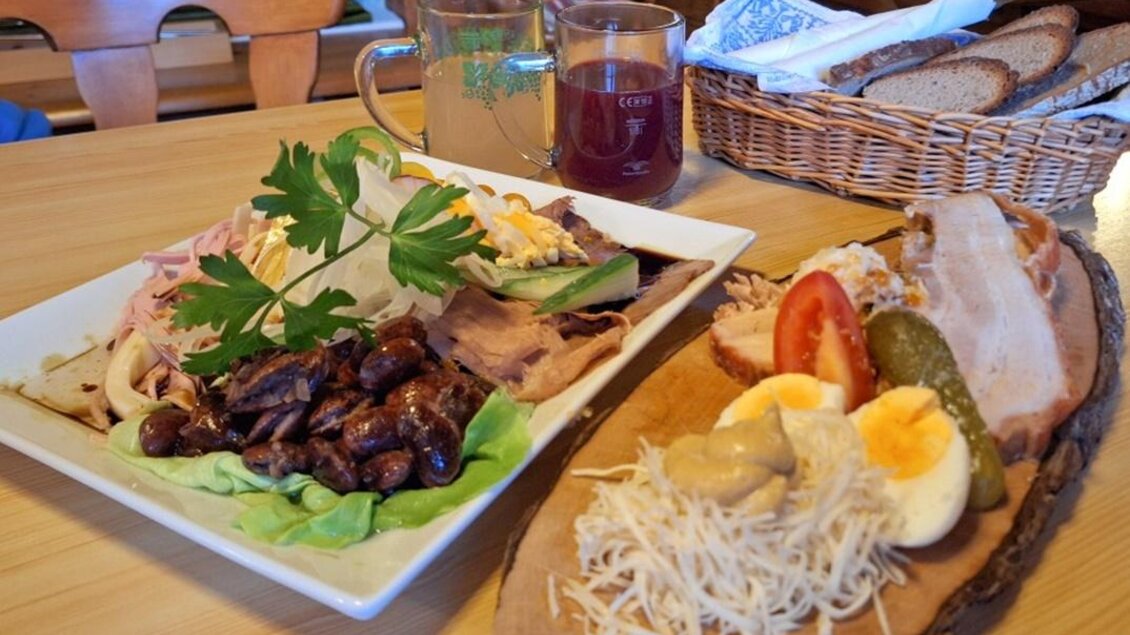 Ein appetitliches Tellergericht mit Fleisch, Salat und Gemüse auf einem Holzbrett. Im Hintergrund sind Getränke und ein Korb mit Brot zu sehen. | © WEGES