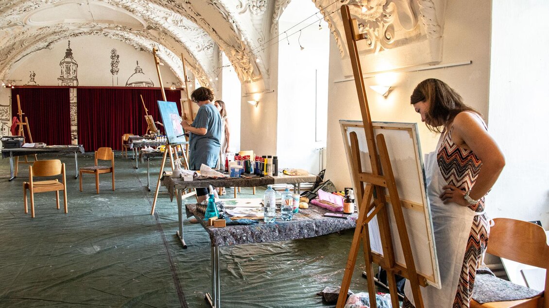 Ein kreativer Raum mit mehreren Malerarbeiten und Staffeleien. Zwei Künstlerinnen arbeiten an ihren Bildern in einer hellen Umgebung.