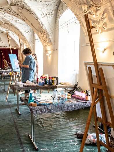 Ein kreativer Raum mit mehreren Malerarbeiten und Staffeleien. Zwei Künstlerinnen arbeiten an ihren Bildern in einer hellen Umgebung.