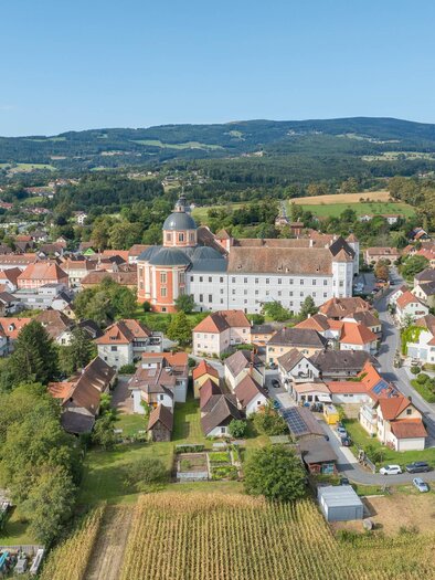 A picturesque view of a small village with colorful houses and an impressive monastery. The surrounding landscape is green and hilly. | © Helmut Schweighofer