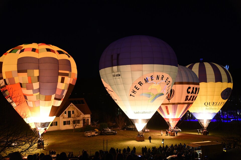 Bunte Heißluftballons leuchten in der Dunkelheit und ziehen viele Zuschauer an. Im Hintergrund sind ein Haus und eine Menschenmenge zu sehen.