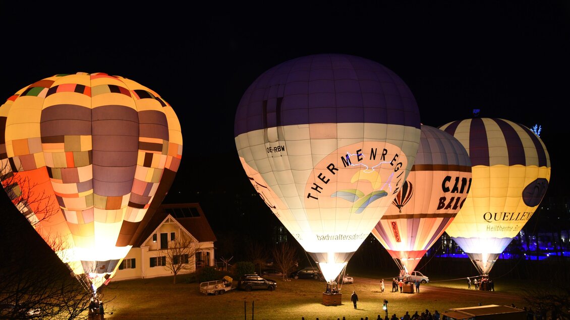 Bunte Heißluftballons leuchten in der Dunkelheit und ziehen viele Zuschauer an. Im Hintergrund sind ein Haus und eine Menschenmenge zu sehen.